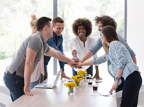 Grroup of six women and men of diverse backgrounds working together. They are 'hands in' across a conference table with yellow flowers in the center and some folders and laptops.